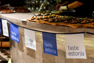 Eesti stiilis suupisted välismaa messil. Suupistetelaua taga ripub lint lipukestega, millel kiri "Taste Estonia".